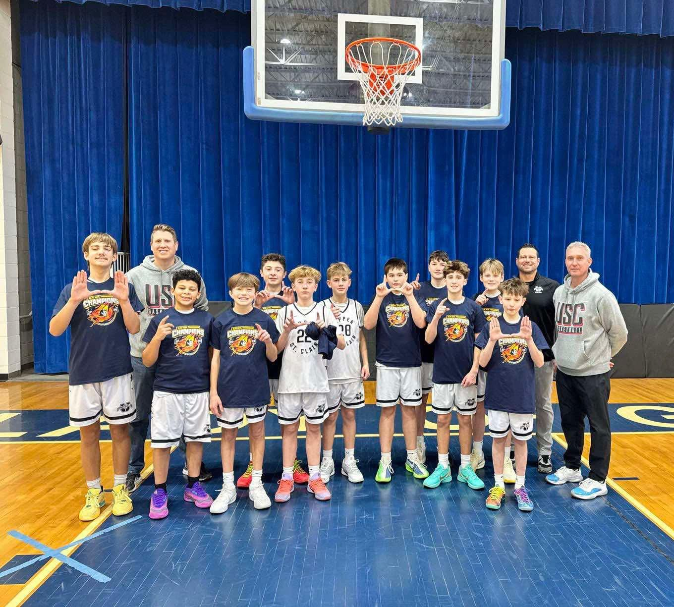 Youth basketball team posing on a court under a hoop, wearing navy jerseys with a coach at each end.