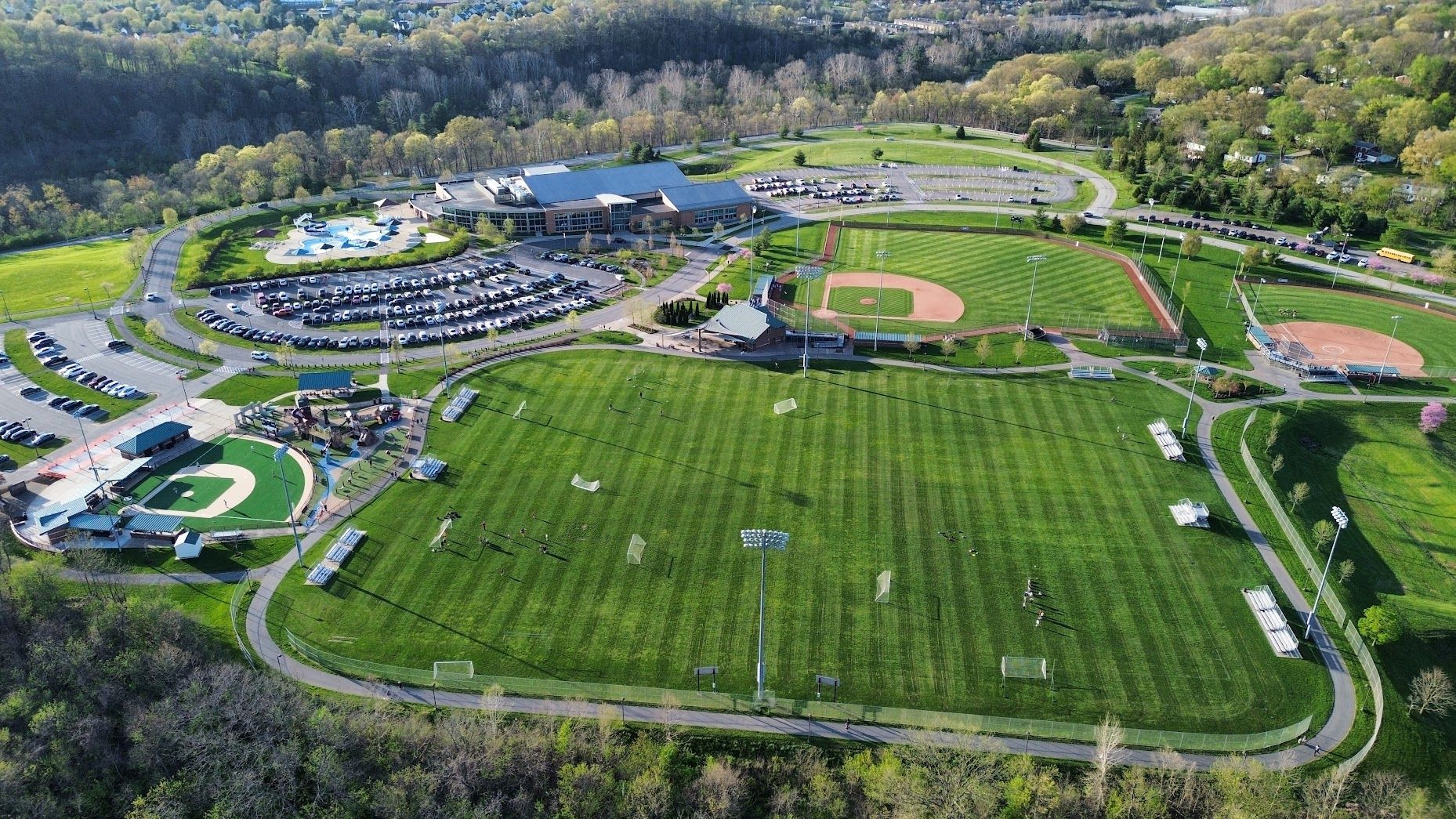 Aerial view of a baseball field and large green sports complex surrounded by trees.