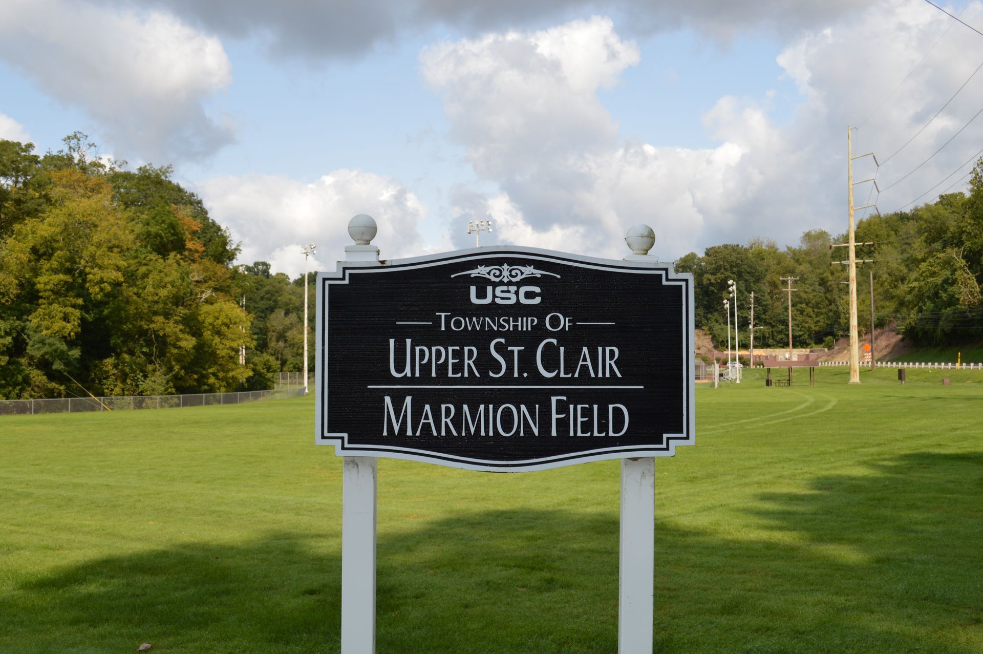 Town sign for Upper St. Clair, Marion Field, on a grassy field with trees and cloudy sky.