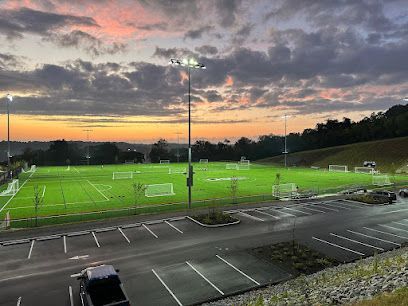 Soccer field at sunset with bright floodlights, parking lot in foreground, and dramatic cloudy sky