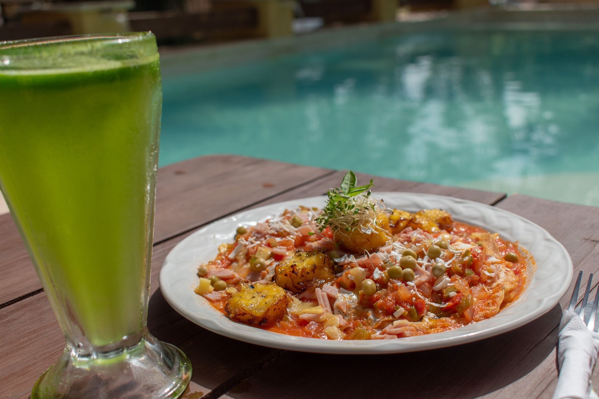 Un plato de comida y un vaso de jugo verde están en una mesa al lado de una piscina.