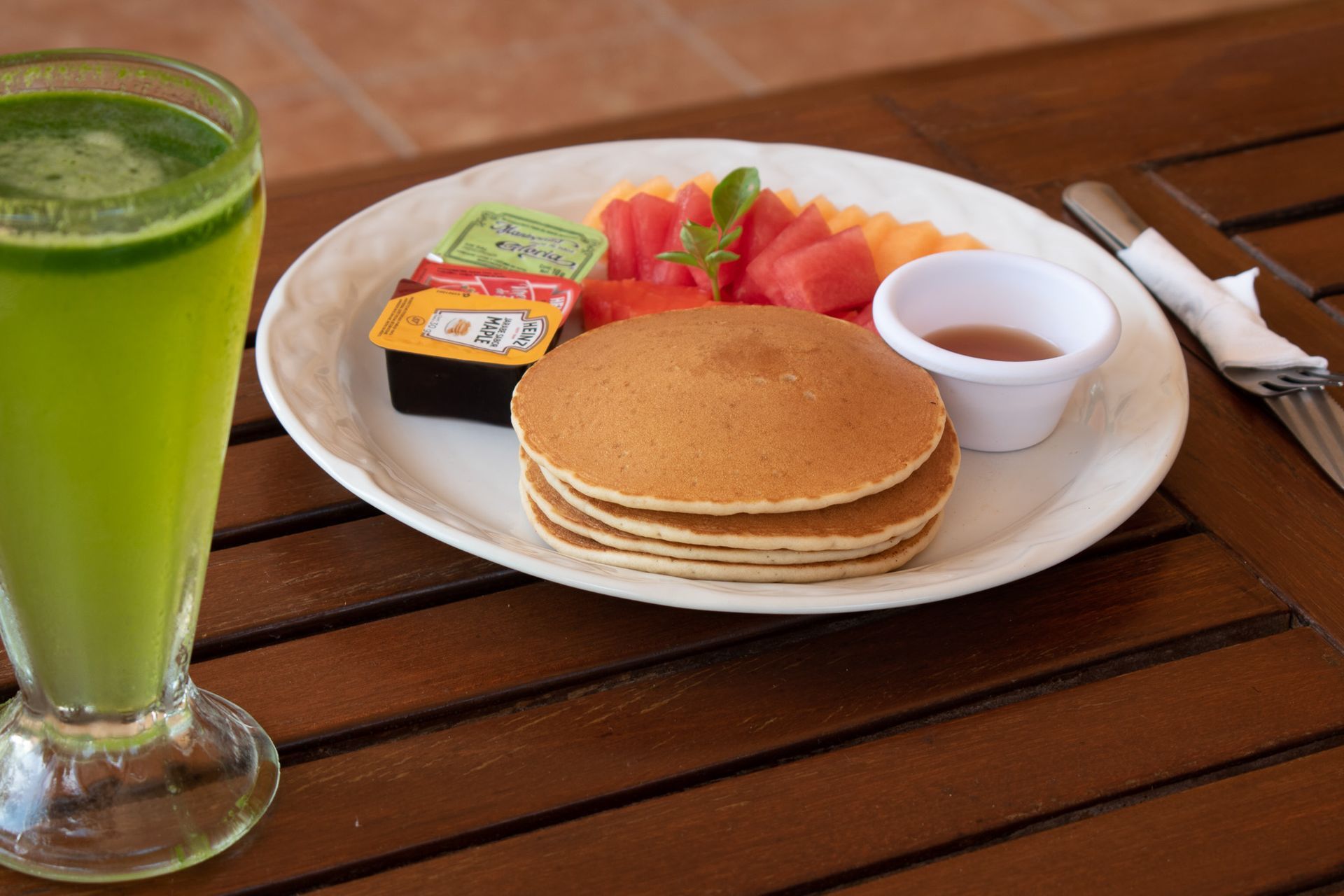 Un plato de panqueques y fruta junto a un vaso de jugo verde en una mesa de madera.