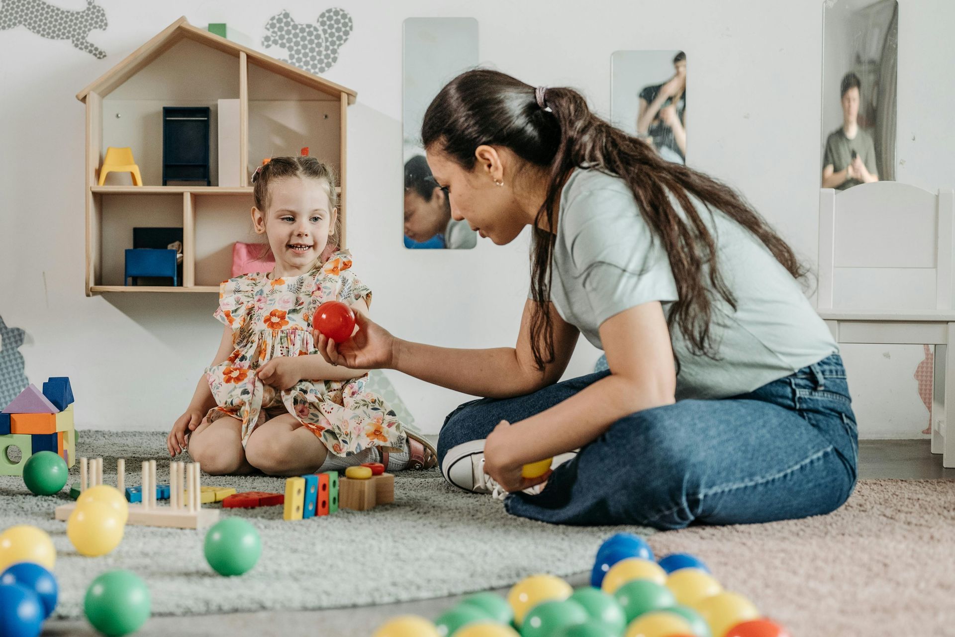 Woman and child playing with colorful balls on a rug in a playroom, looking at each other with smiles.