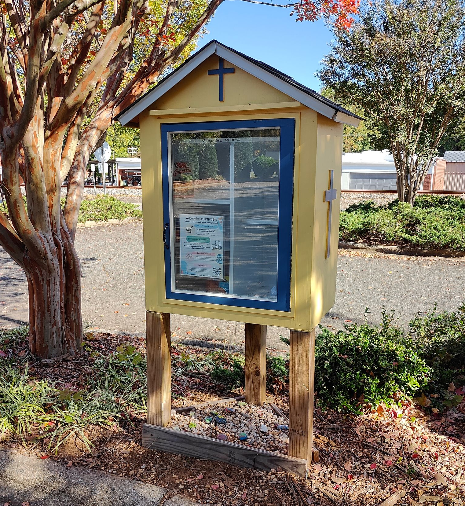 Yellow community food pantry with blue trim, cross on the roof, raised on wooden posts.