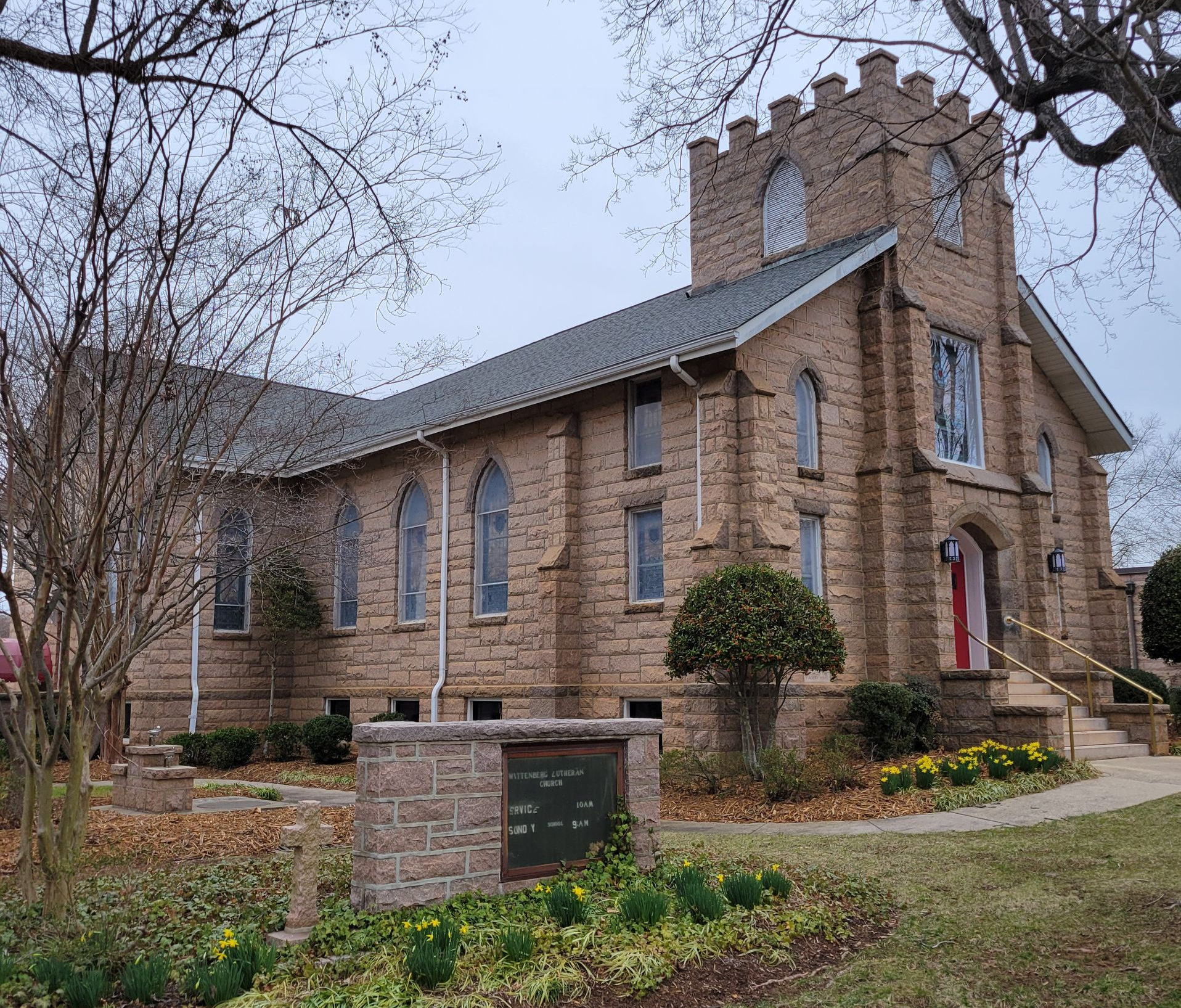 Night view of the church lit by luminaries lining the sidewalk and a Christmas wreath on the red door.