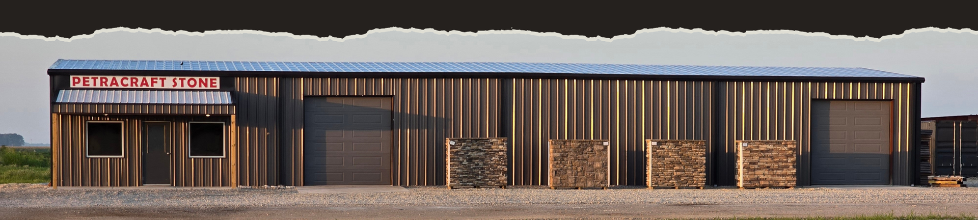 A long, dark-brown metal industrial building with a storefront entrance, two roll-up doors, and stacks of lumber outside.