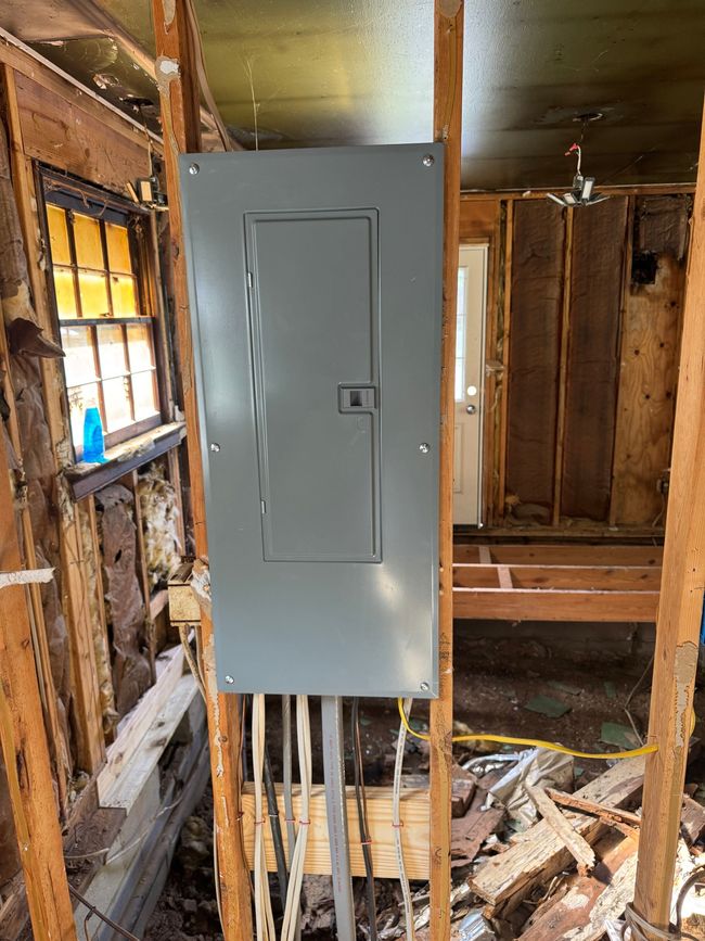 Gray electrical panel mounted on exposed wall framing in a damaged, unfinished room