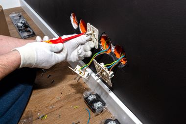Gloved hands wiring a small electrical component on a wall with exposed flames from a fire damage test