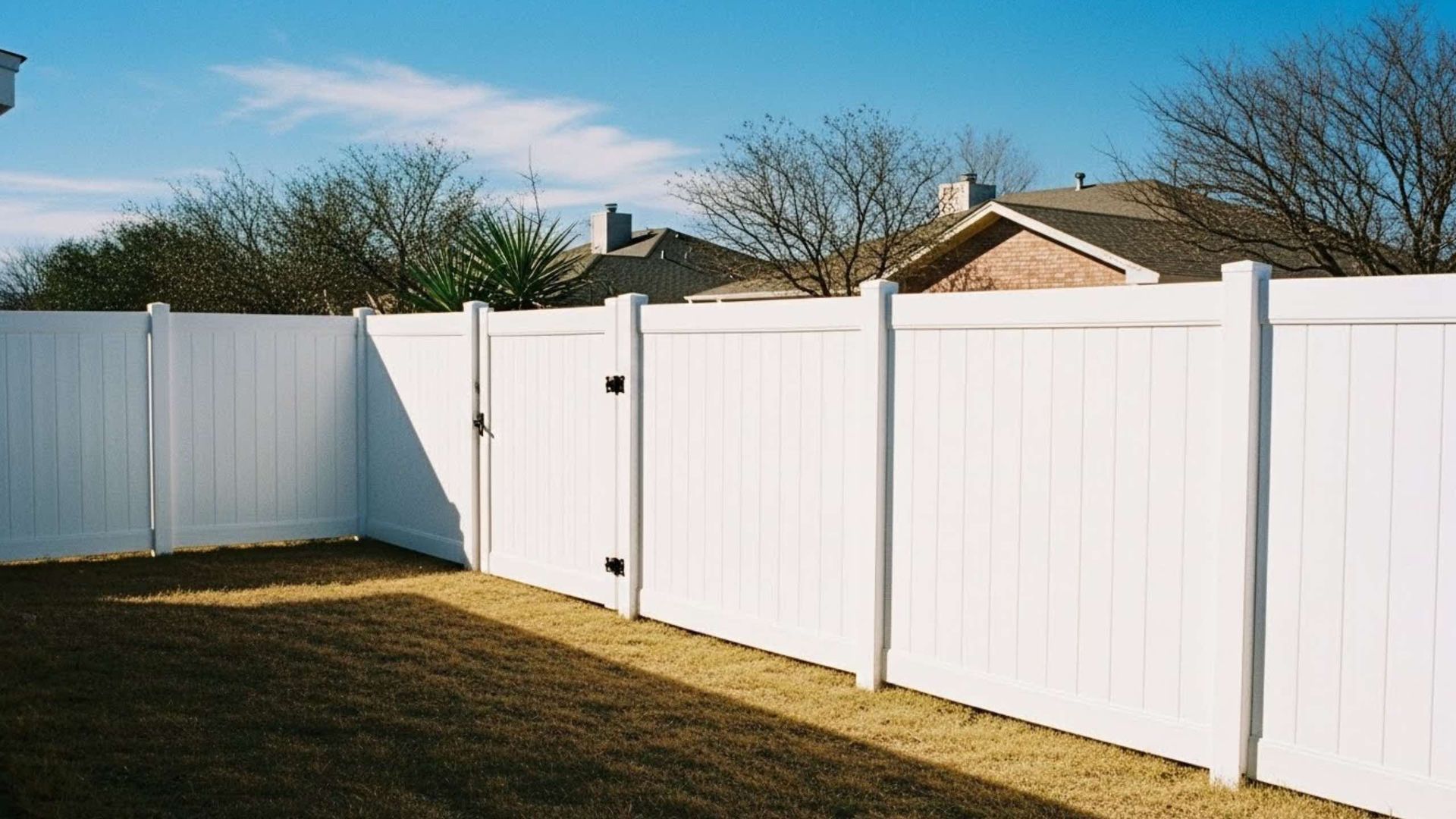 A white vinyl privacy fence encloses a backyard with dry grass under a blue sky.