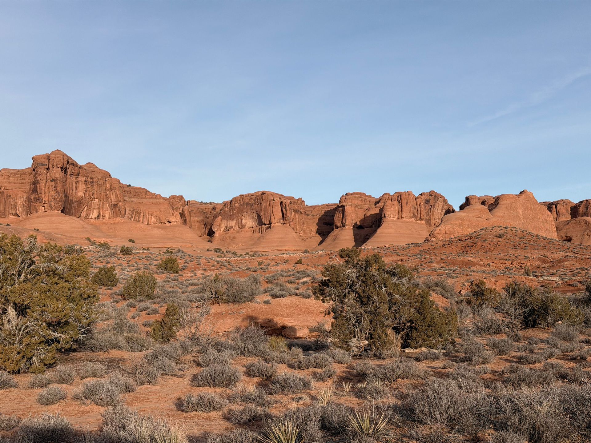 Red sandstone cliffs under a clear blue sky in a desert landscape with scrubby brush.