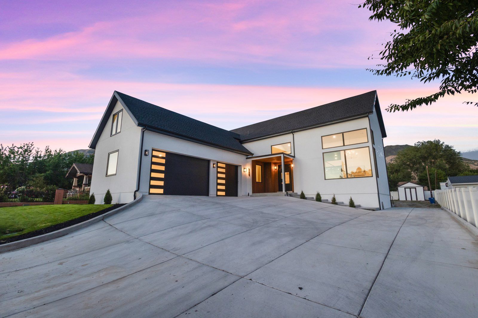 Modern white house with dark roof and garage, concrete driveway, dusk sky.
