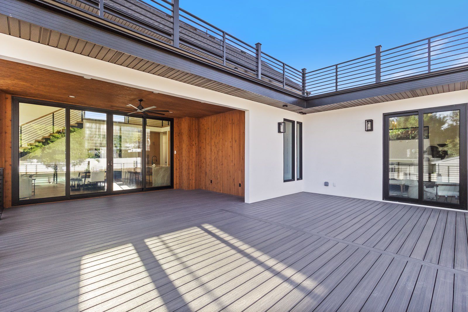 Patio with gray decking, sliding glass doors, wooden accents, and a rooftop deck with metal railing.