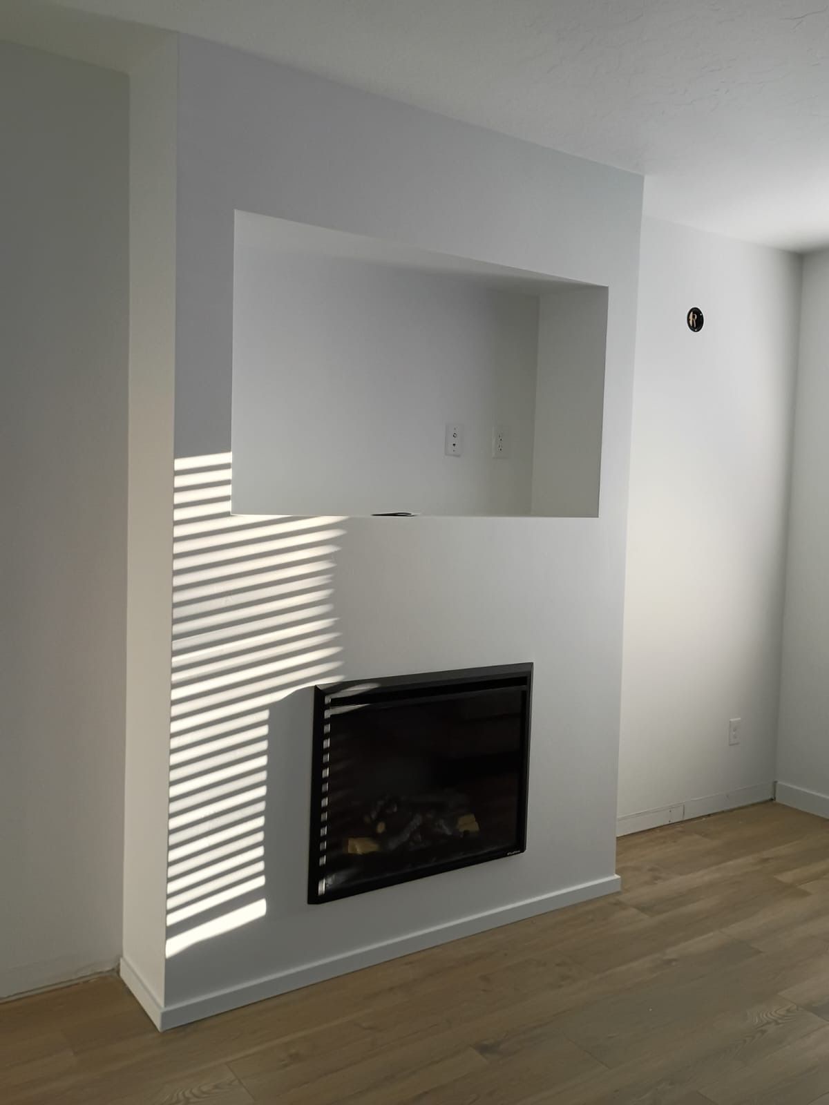 White wall with fireplace and recessed area above. Wooden floor, blinds casting shadows.