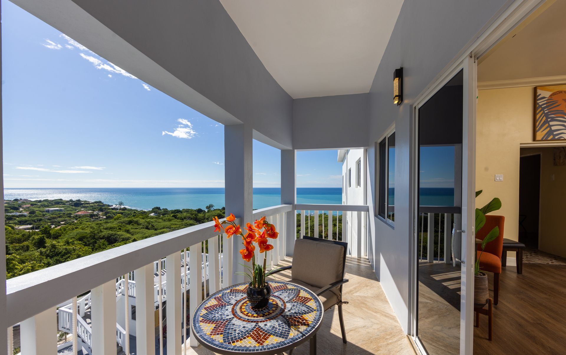 Balcony with sea view. Table with flowers, chair. Blue sky, ocean, and trees.