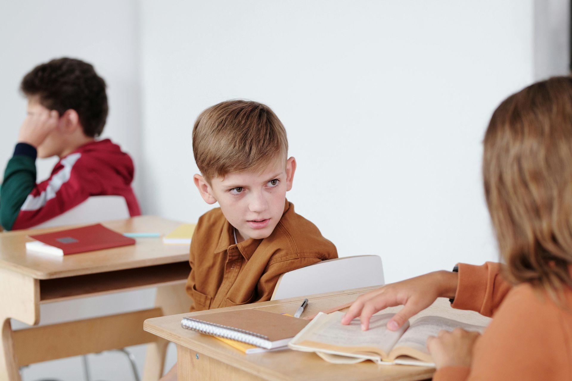 Students sit at desks in a classroom; one boy looks toward a peer who is reading from an open book.