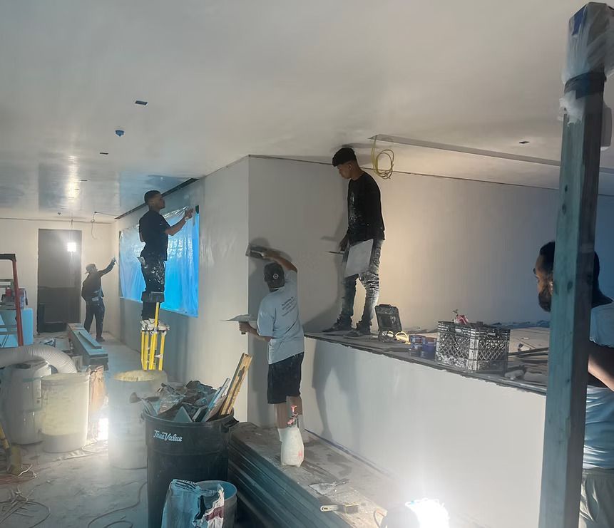 Workers painting a room, with one on a ladder and others on a raised ledge.
