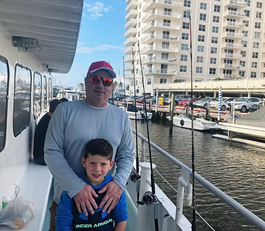 Man in red cap standing with a smiling child on a boat dock by a marina and apartment buildings