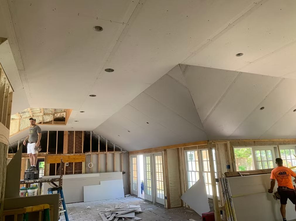 Unfinished room with vaulted white ceiling, exposed framing, and a worker in an orange shirt near the windows