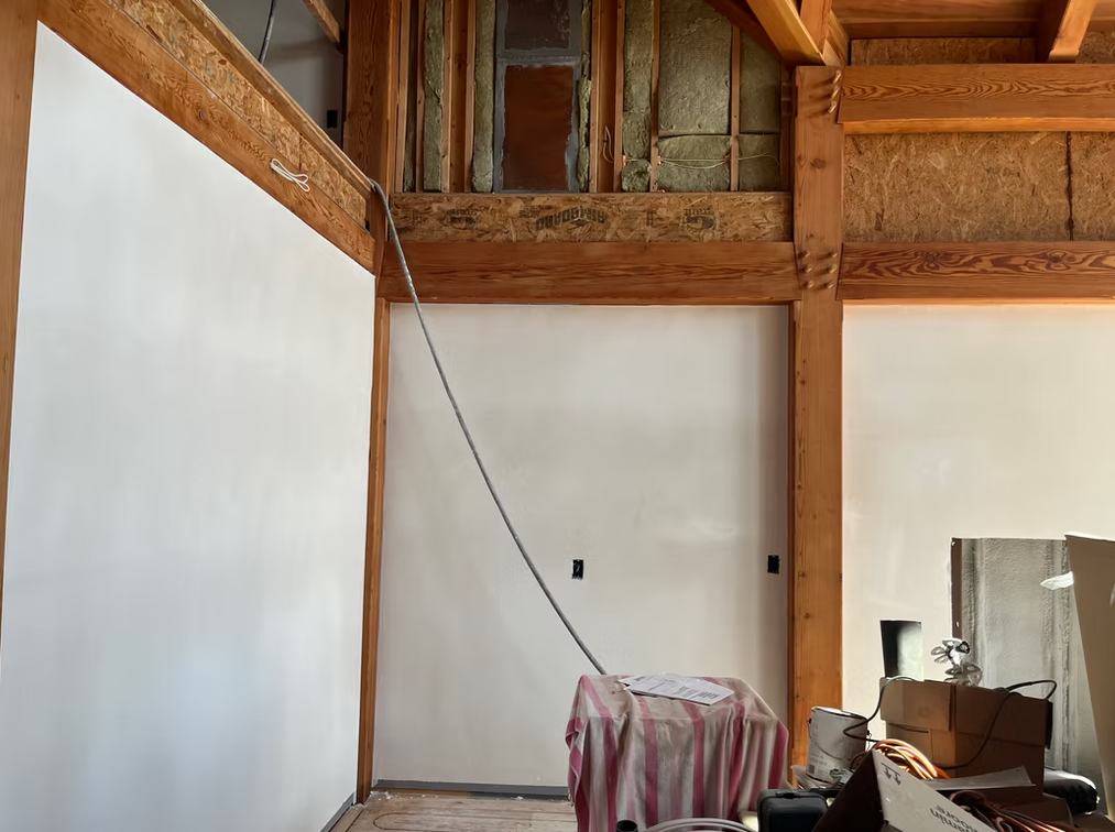 Partially finished room with white walls, wooden beams, hanging cable, and covered chair near the window.