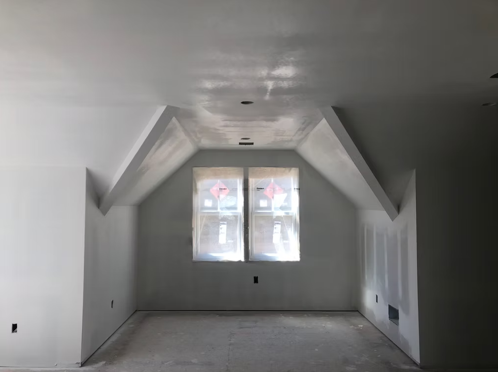 Empty unfinished attic room with white walls, sloped ceilings, and two windows at the far end.