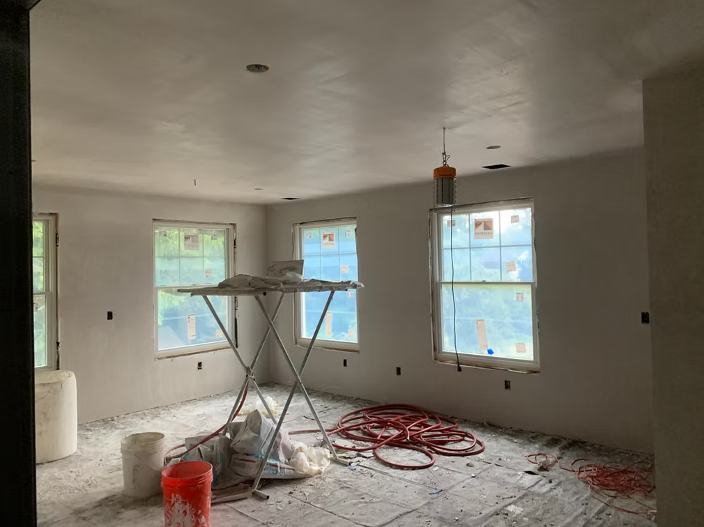 Unfinished room with white walls, three windows, paint-splattered floor, and construction tools centered on a tarp.
