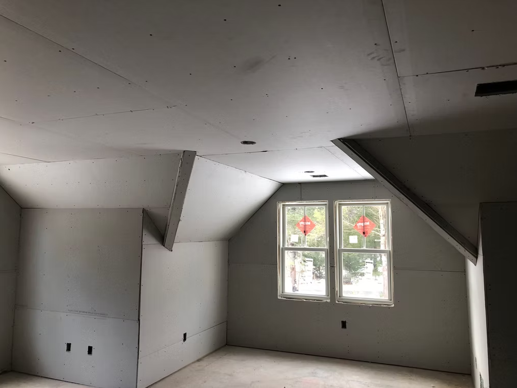 Empty unfinished attic room with sloped drywall ceiling and a small window with flowers outside