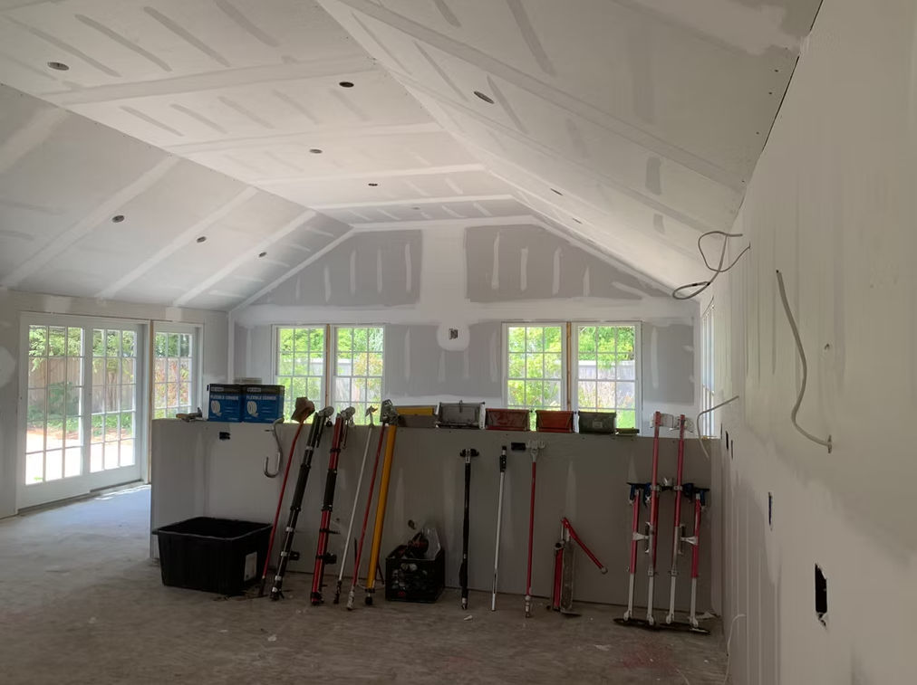 Bright unfinished room with vaulted white ceiling, large windows, and tools leaning against a wall