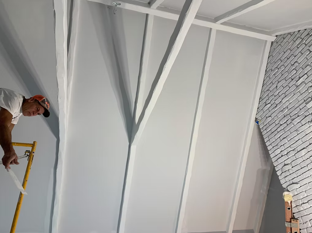 Worker painting white ceiling beams indoors, with a ladder and patterned wall visible