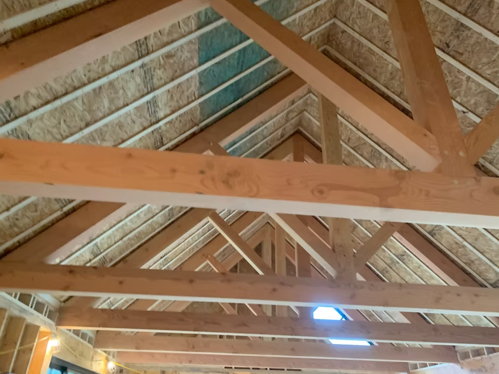 Exposed wooden roof trusses inside an attic, with slanted beams and a small bright skylight opening.