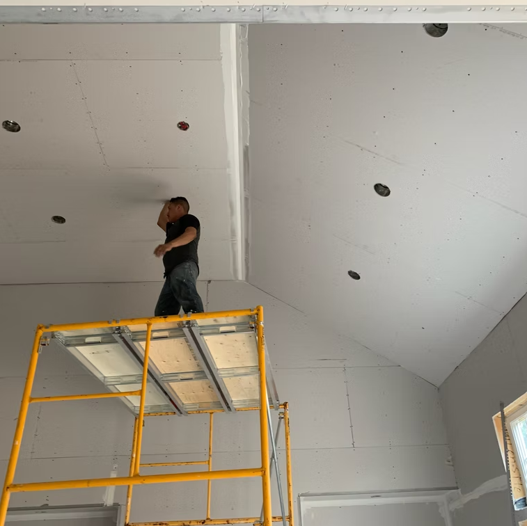 Worker standing on a yellow scaffold, inspecting a white ceiling during interior construction.