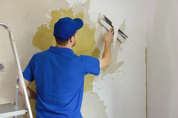 Person in a blue shirt and cap painting a wall with a putty knife next to a ladder