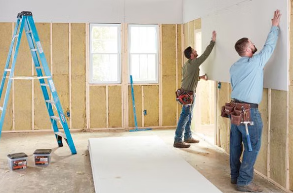 Workers installing drywall in a room under renovation, with a blue ladder and tools on the floor