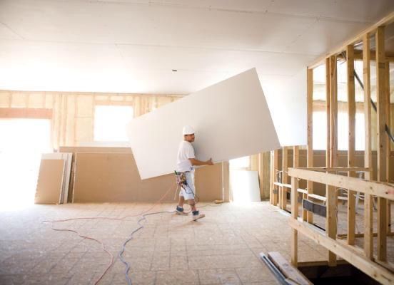 Worker carrying a large drywall sheet inside a room under construction
