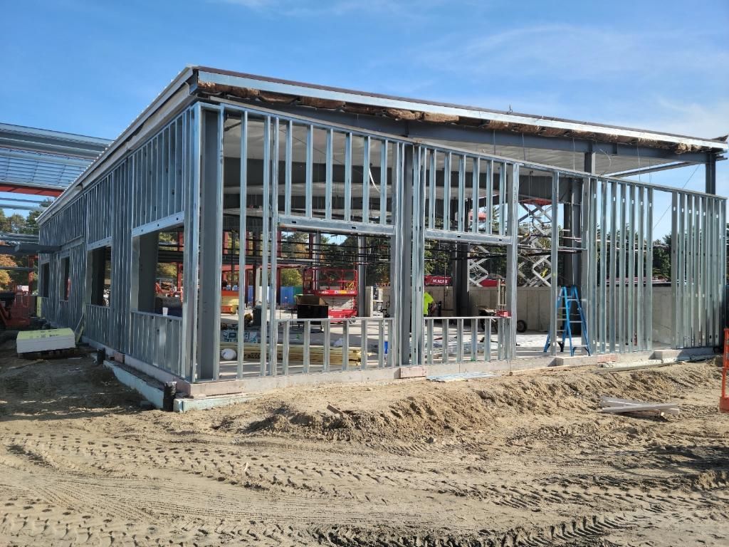 Steel-framed building under construction on a dirt site, with exposed studs and open window and door frames.