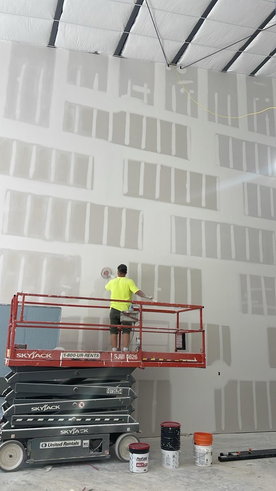 Worker on a red lift applying drywall mud to a large white interior wall in a construction site