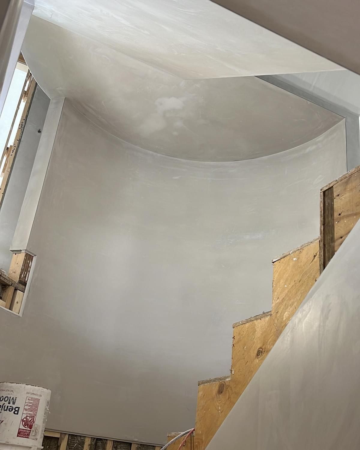 Curved stairwell with white walls and a skylight, showing exposed wooden steps and unfinished construction.