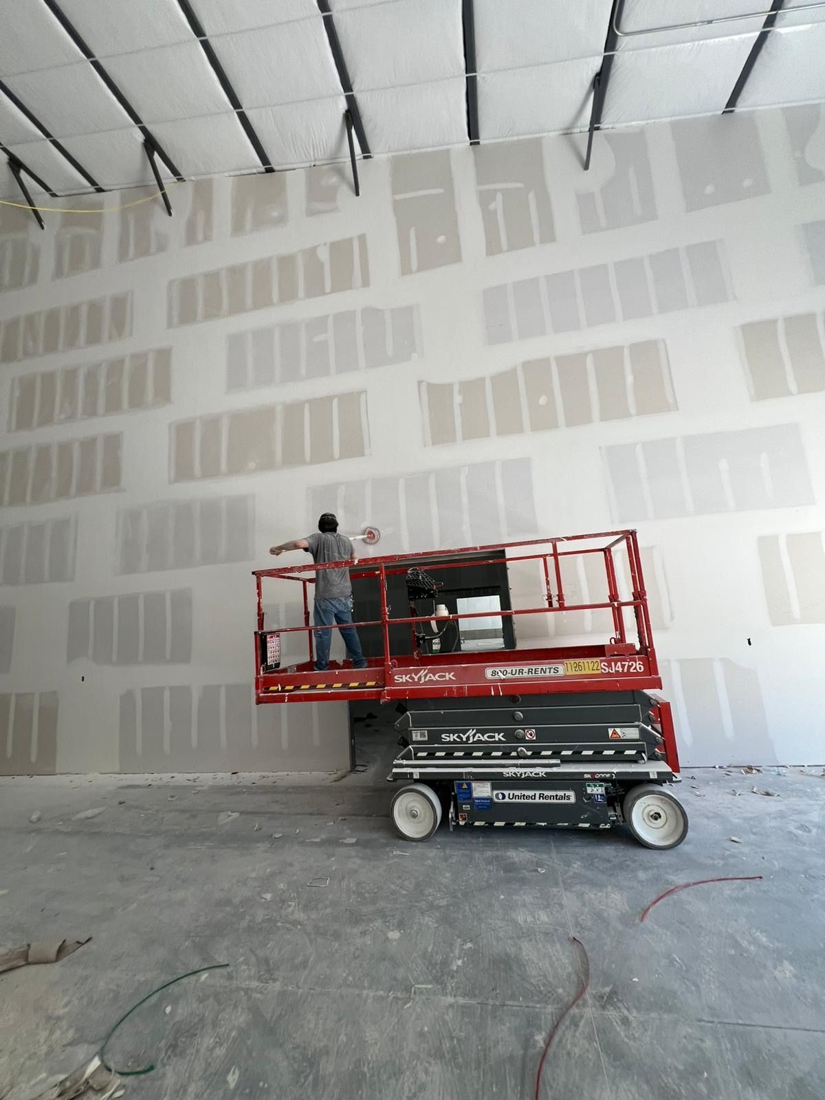 Worker on a red lift near a large drywall wall inside a construction site.