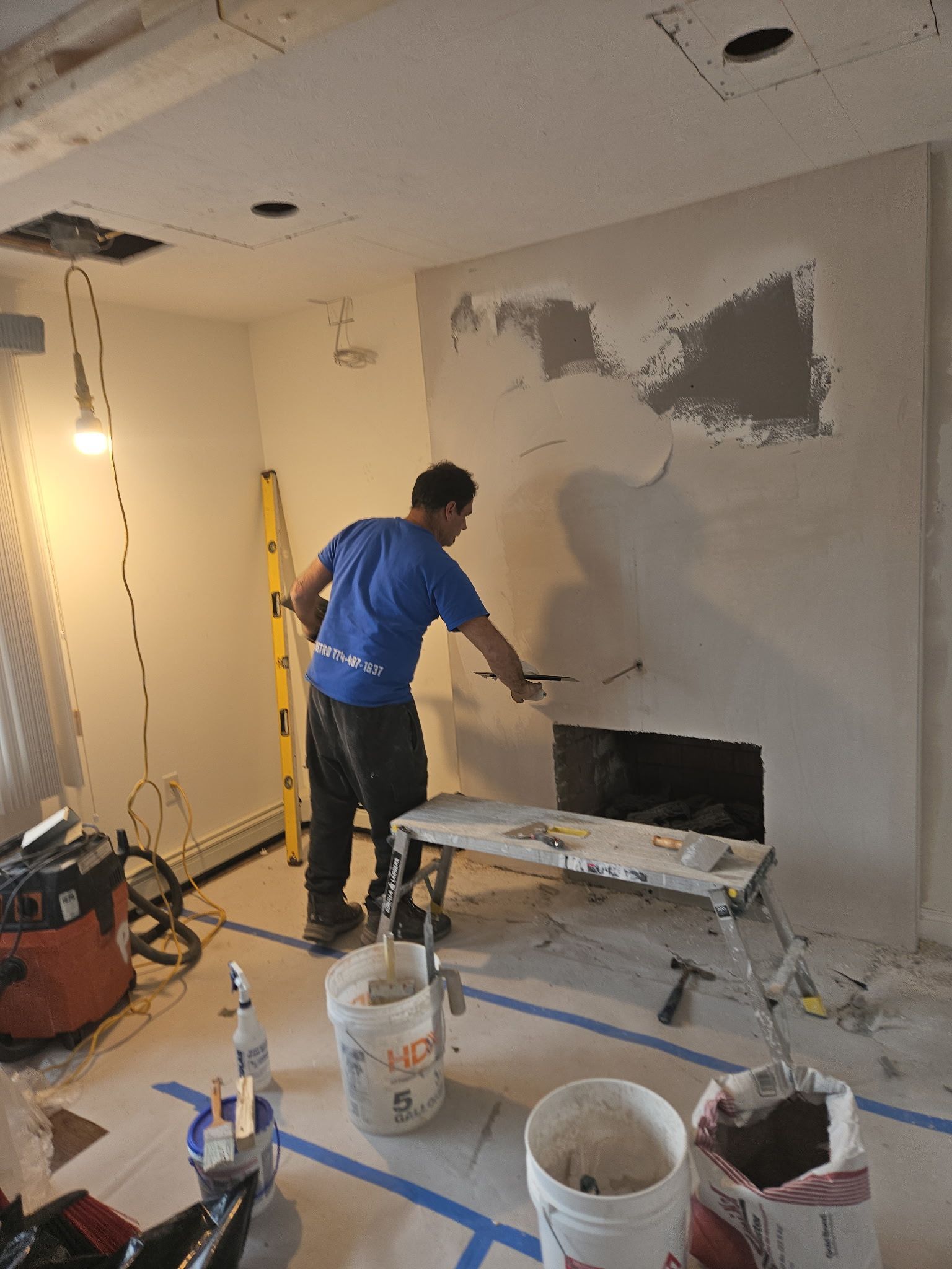 Person plastering a wall in a room under renovation, with tools and paint supplies on the floor.