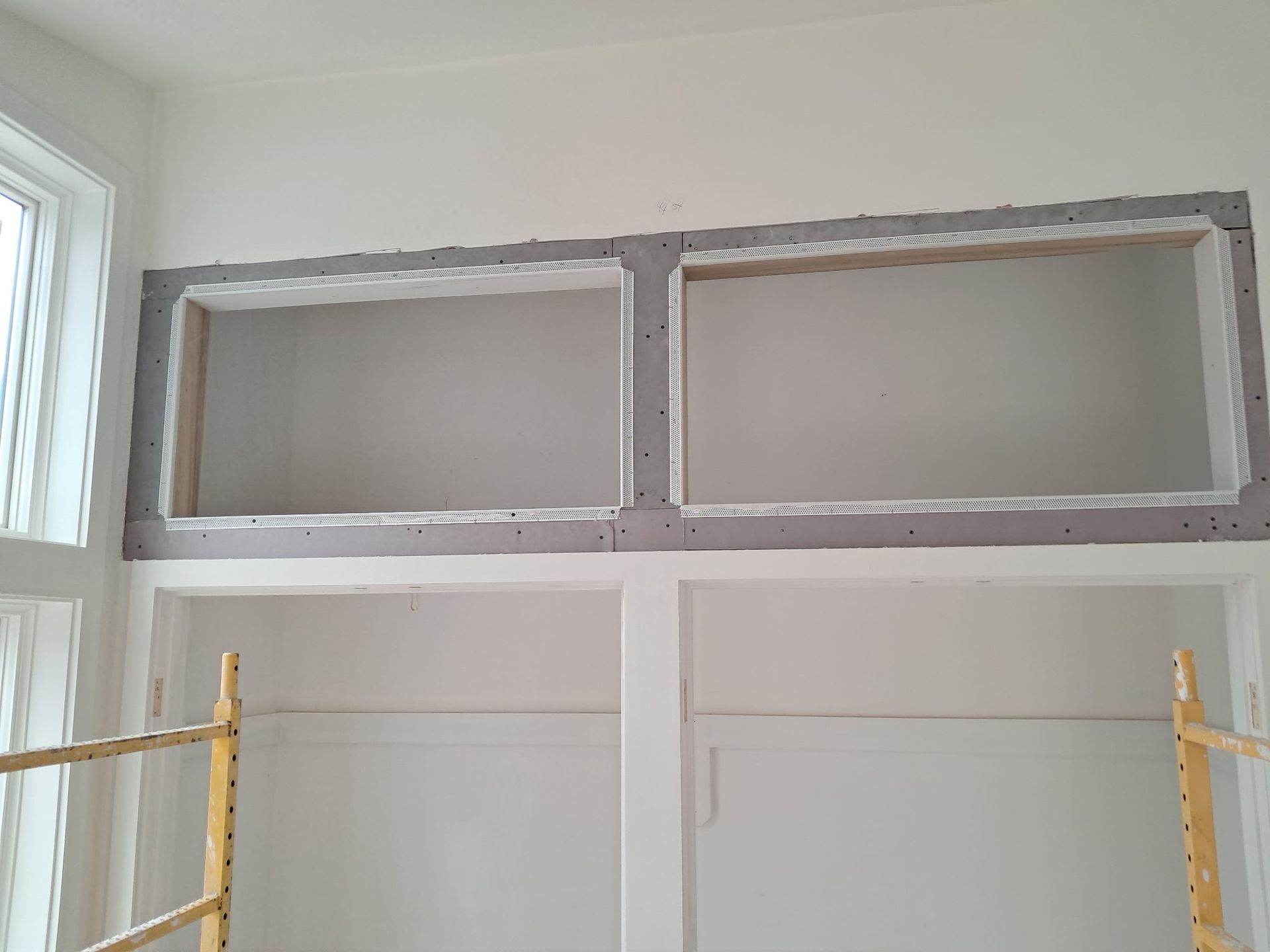 Unfinished wall with two open rectangular cutouts above white doors, framed by yellow scaffolding.
