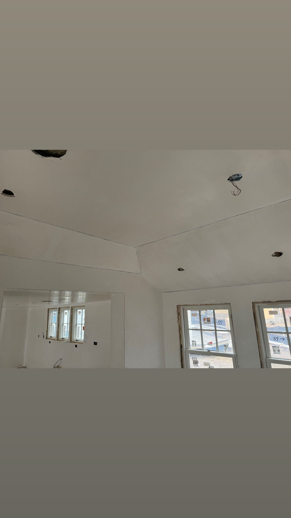 White room under construction with recessed ceiling openings and windows along the wall.
