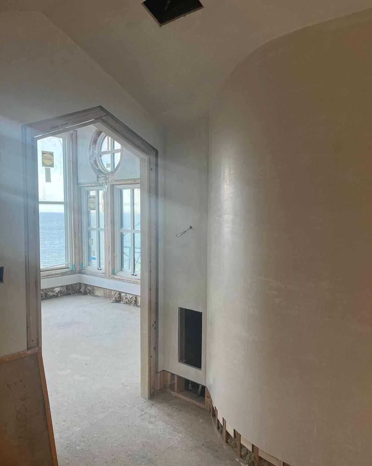 Empty hallway opening into a bright room with large bay windows and unfinished walls
