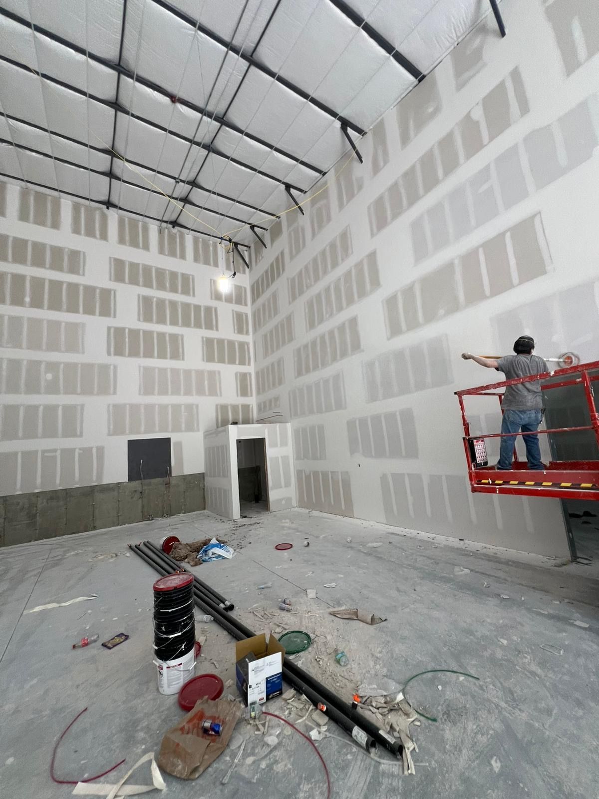 Large unfinished warehouse interior with white drywall, scattered tools, and a worker on a red lift near the right wall