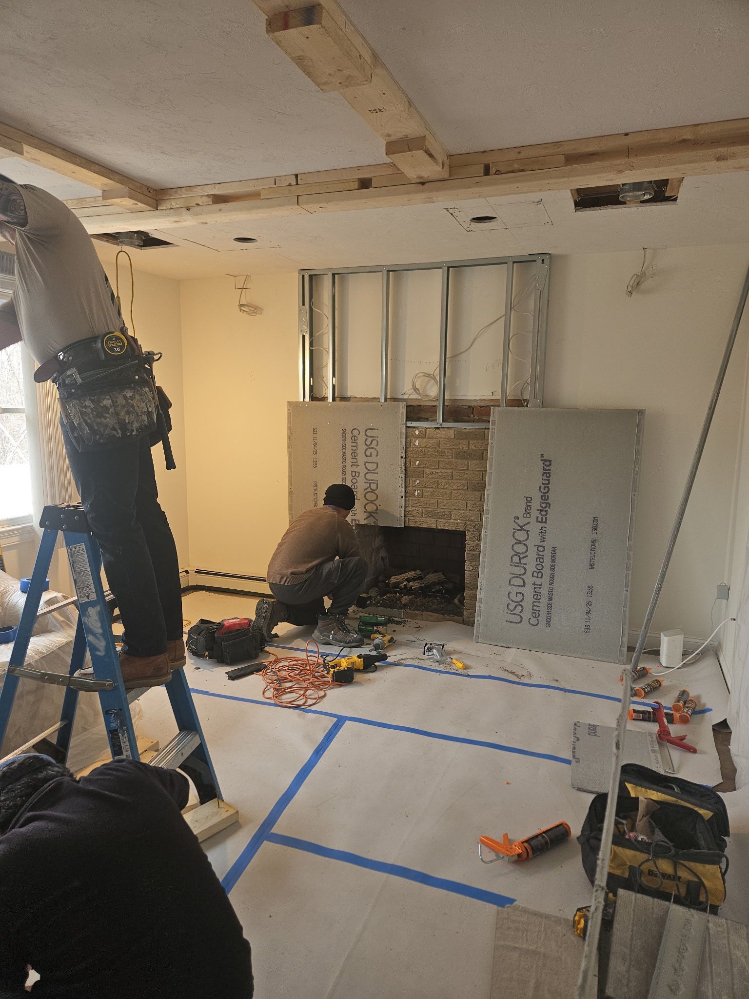 Worker kneeling by fireplace in a room under renovation, with blue tape on the floor and tools scattered around.