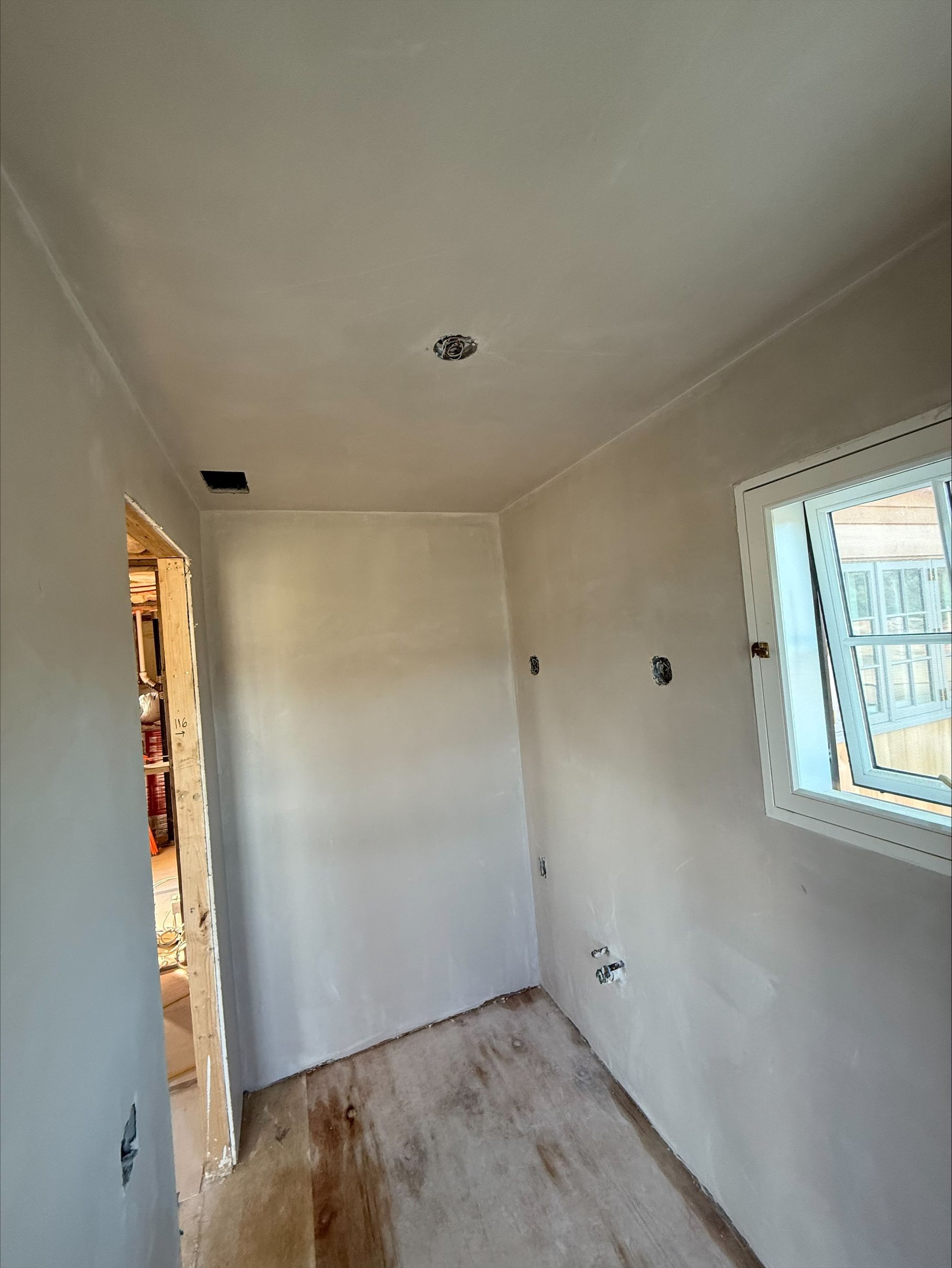 Narrow unfinished room with white walls, bare floor, and a window letting in daylight.