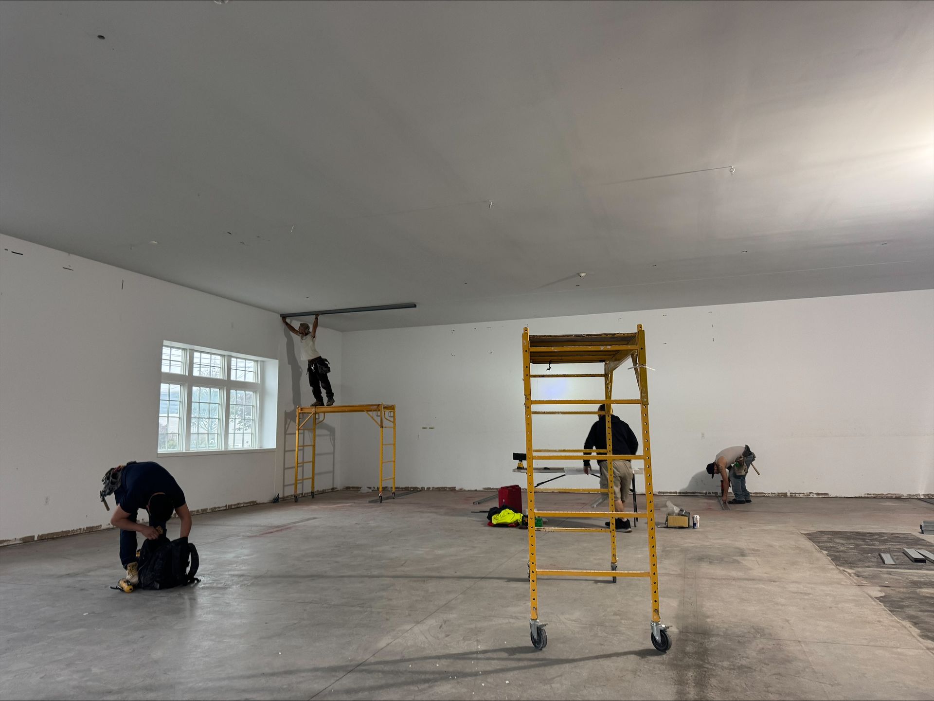 Workers in a large empty room, painting walls and ceiling with ladders and tools.