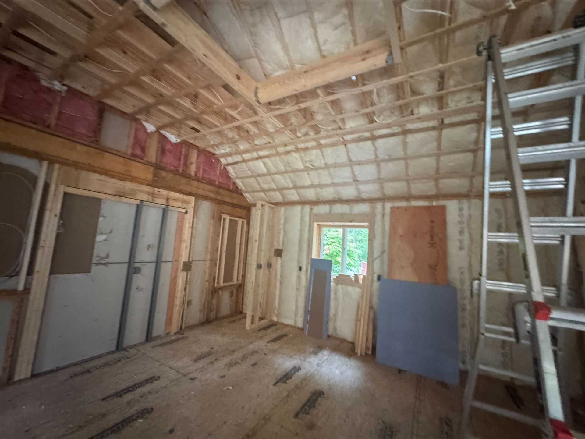 Unfinished room under construction with exposed framing, insulation, ladder, and a window at the far end.