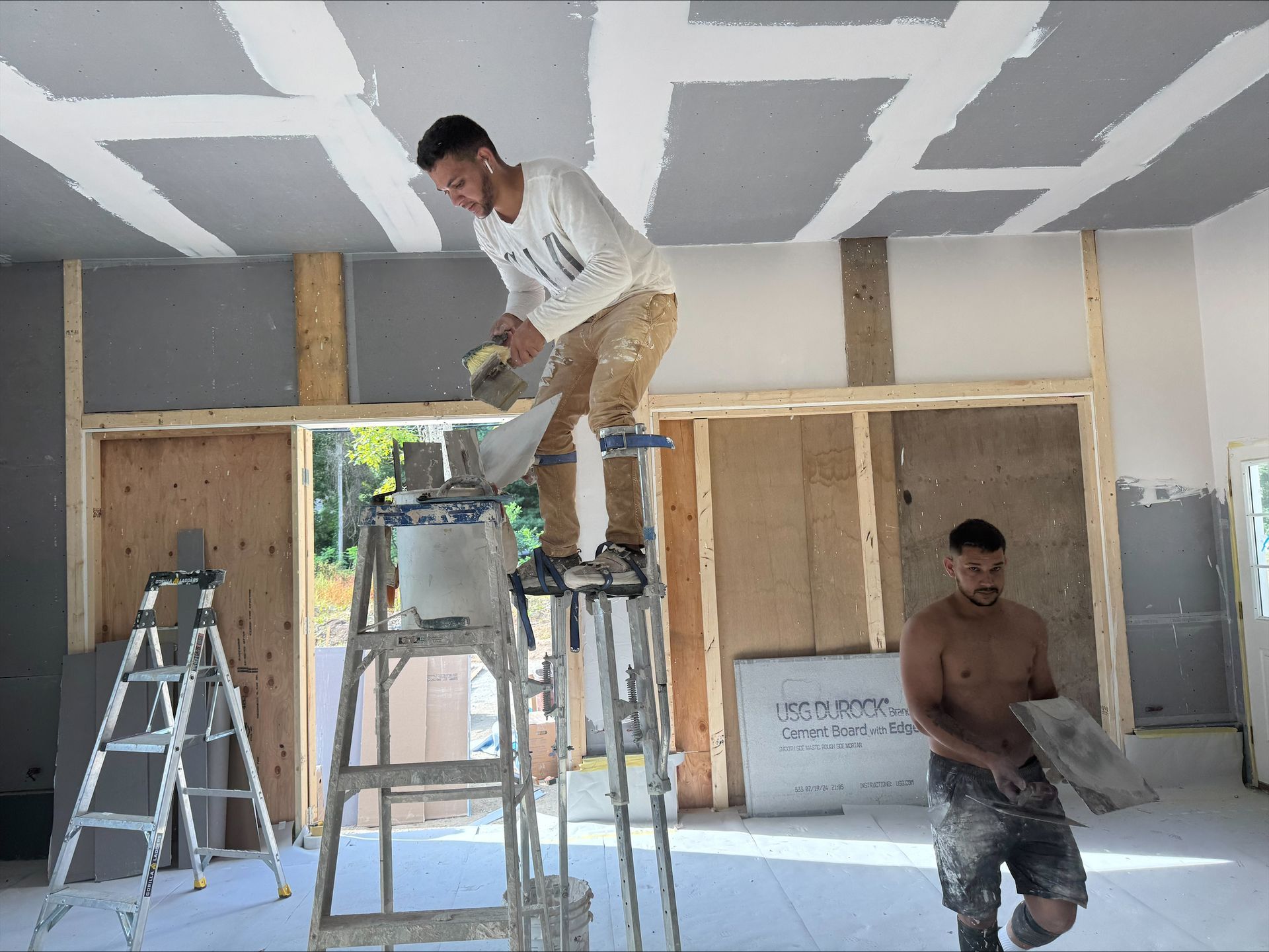 Two construction workers on ladders drywalling an unfinished room with exposed framing.