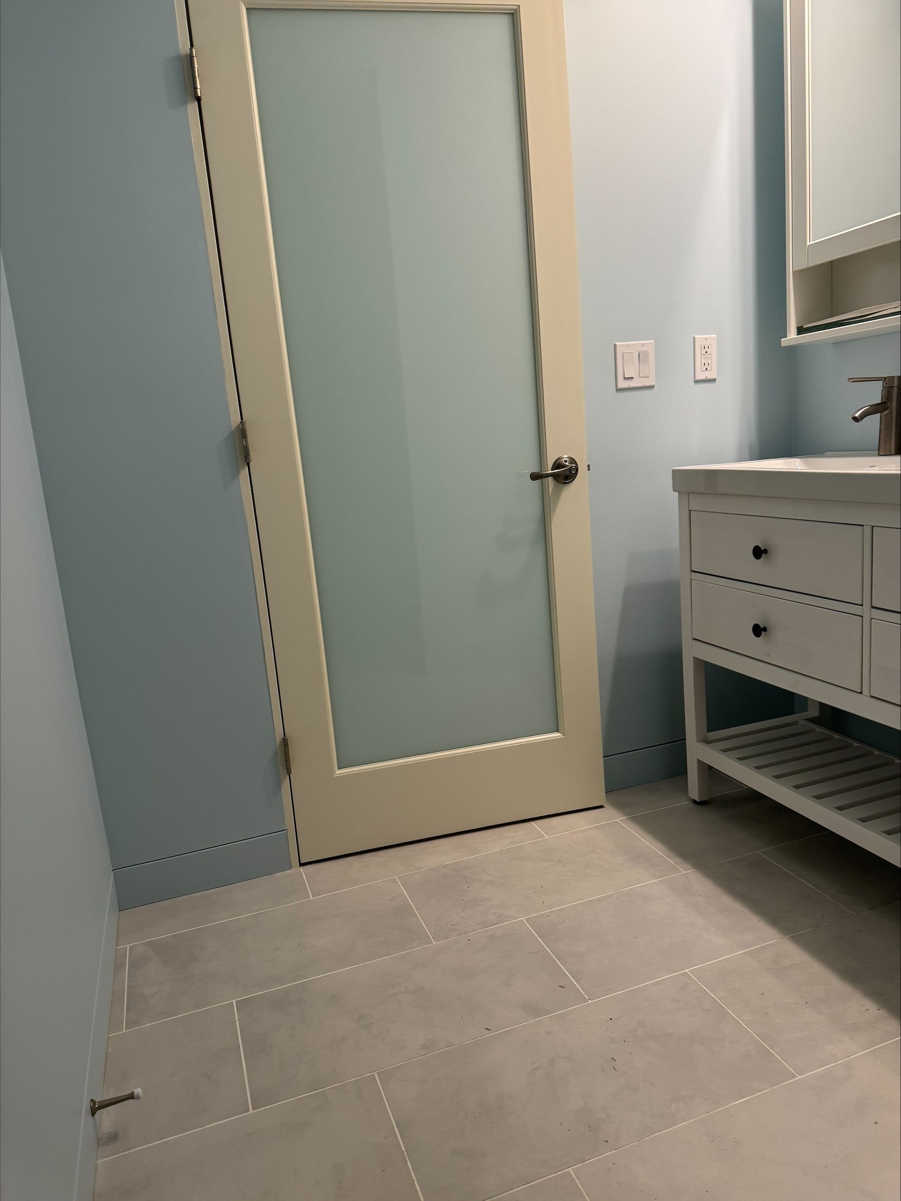 Small bathroom with frosted glass door, light blue walls, tiled floor, and white vanity sink.