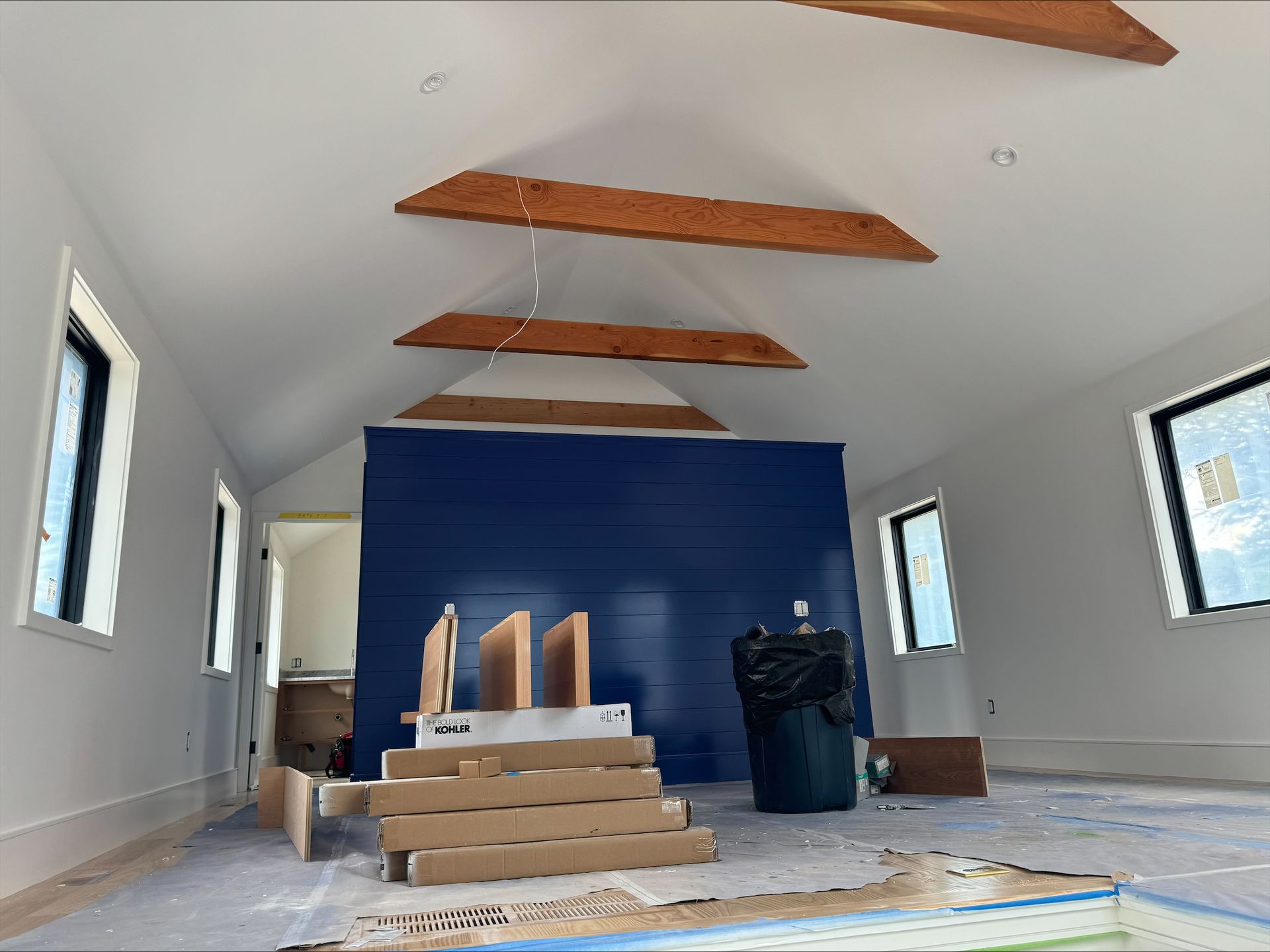 Long room under renovation with blue accent wall, exposed beams, windows, and construction materials.