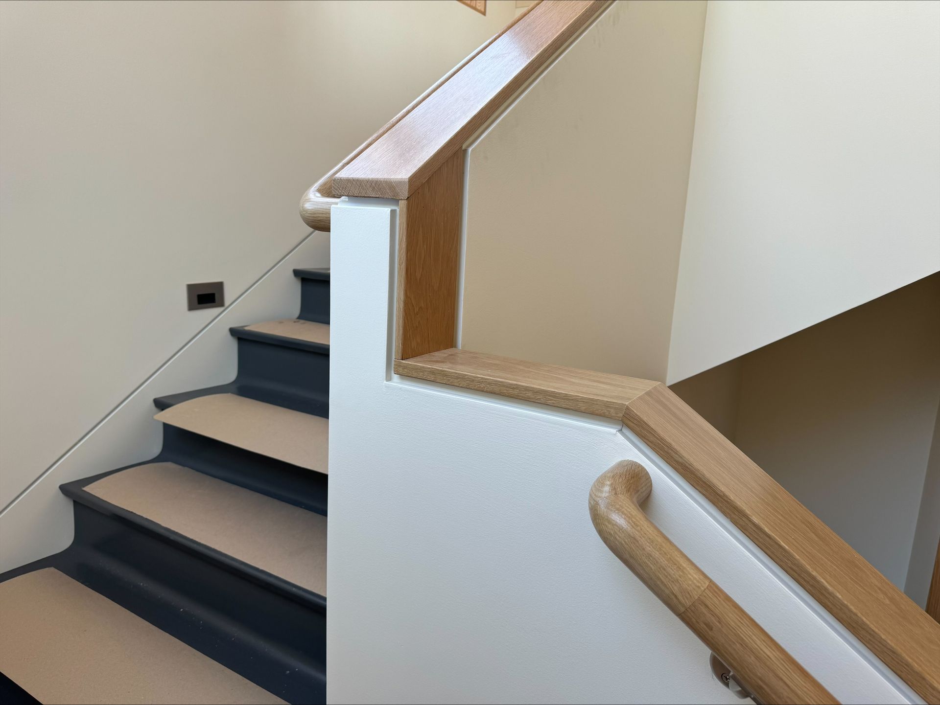 Indoor staircase with dark steps, beige walls, and a light wood handrail around a landing.